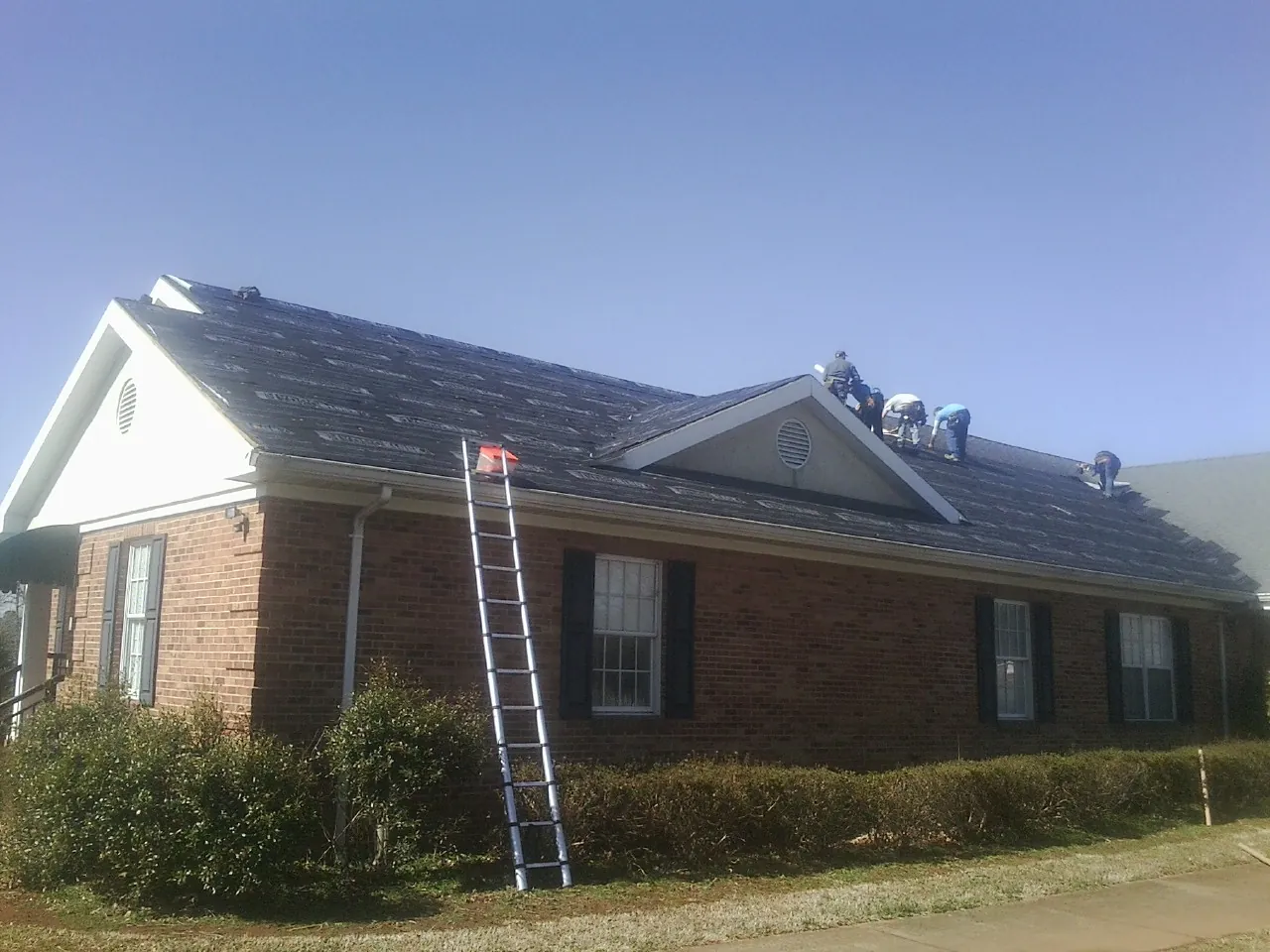Crew tearing shingles off a brick ranch roof with a ladder set — Emergency Roof Repair work by our crew in Anaconda