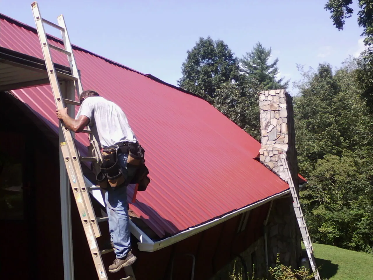 Roofer on a ladder beside a red metal roof and stone chimney — Soffit & Fascia Repair by Roofing Brothers in Anaconda